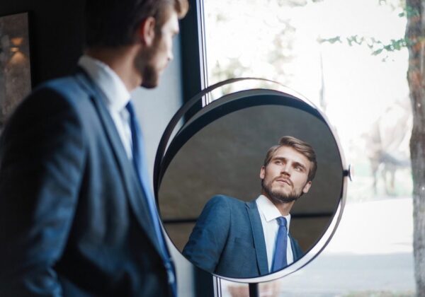 Uomo elegante in giacca e cravatta che si osserva con attenzione allo specchio, simbolo di narcisismo e autocelebrazione. L'immagine rappresenta le caratteristiche del Disturbo Narcisistico di Personalità , tra cui grandiosità e forte attenzione alla propria immagine.